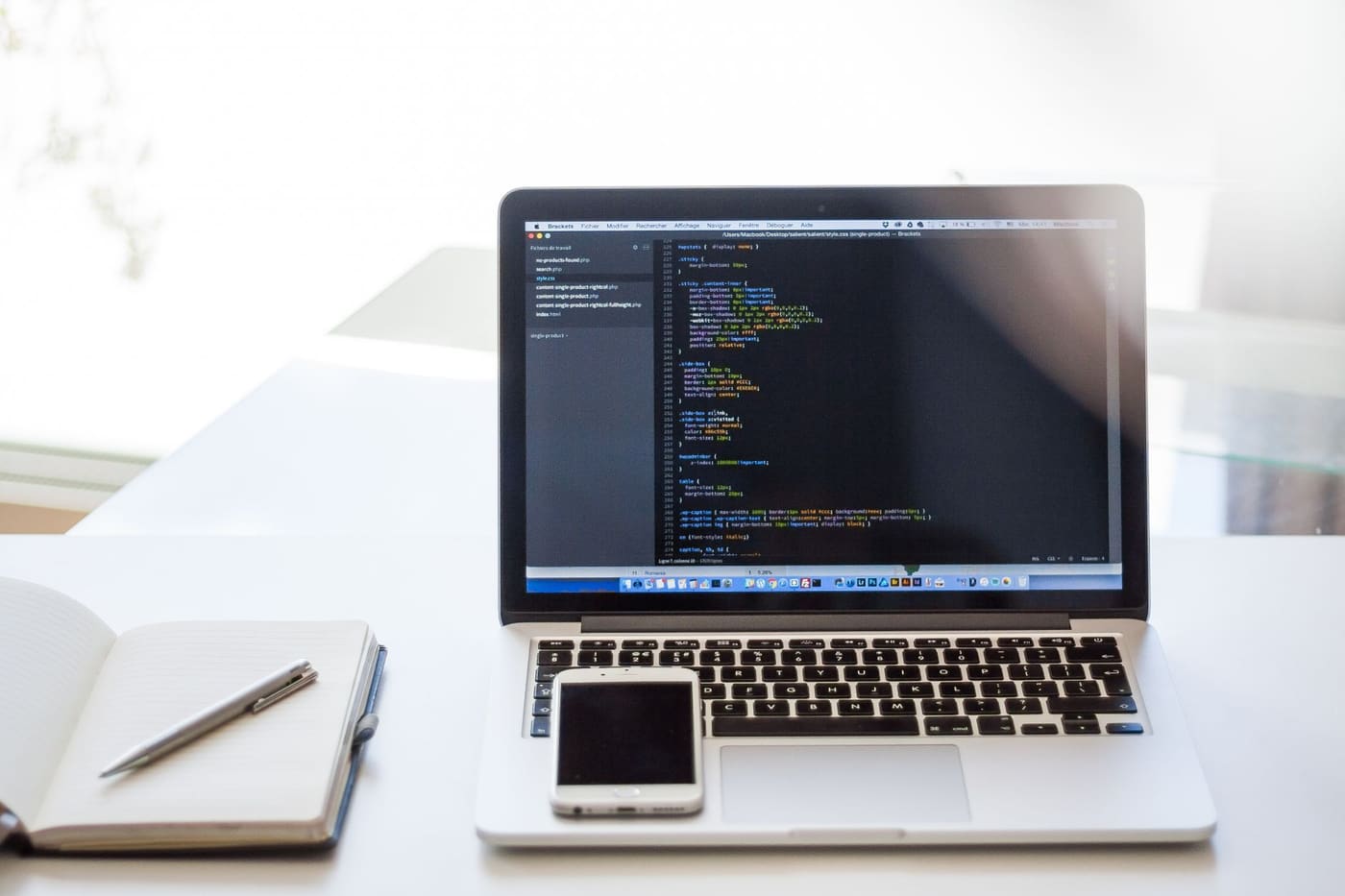 An open laptop showing code on a bright desk beside a notebook, pen and smartphone