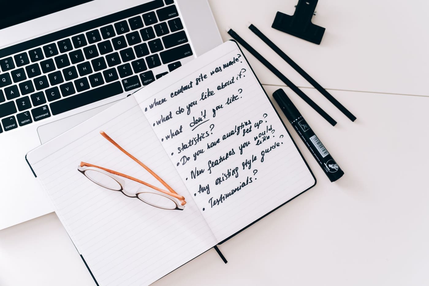 An open notebook with a handwritten list, reading glasses and pens beside a laptop