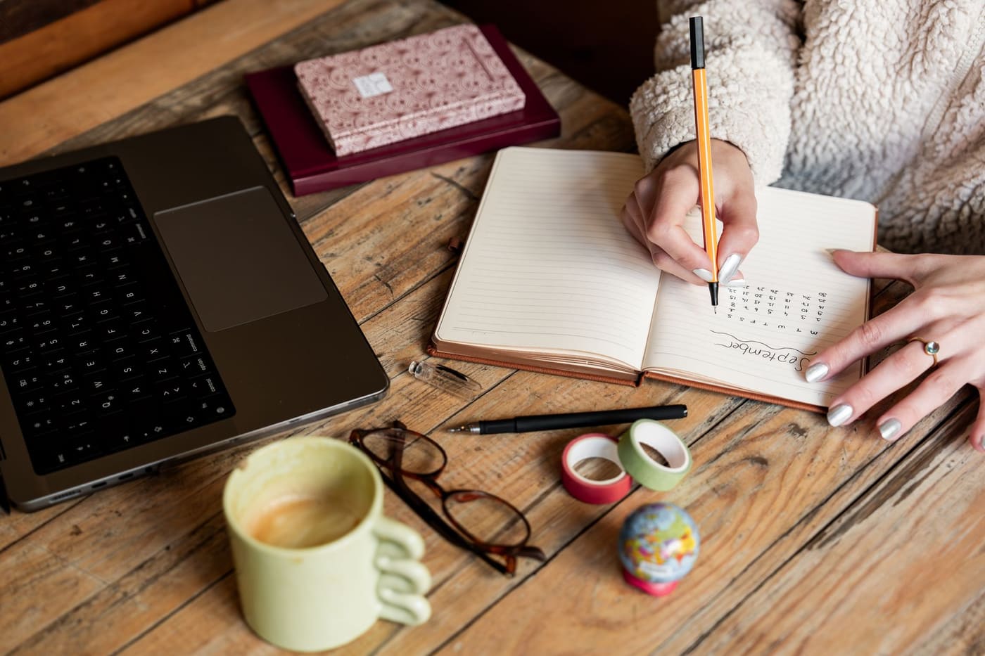 A woman writing a plan in a notebook beside a laptop and a cup of coffee on a wooden desk