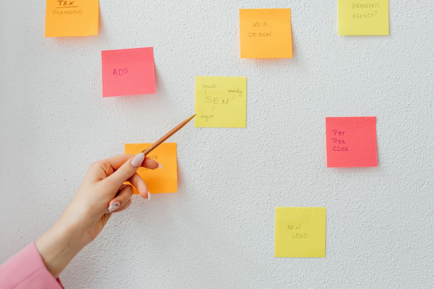 A hand pointing with a pencil at sticky notes arranged on a white wall