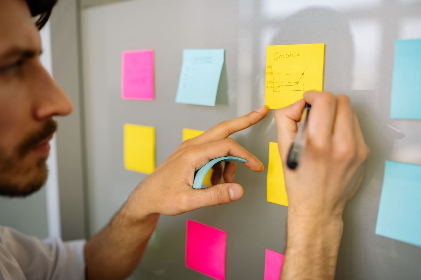 A man in a dark jacket writing on a small sticky note against a light wall